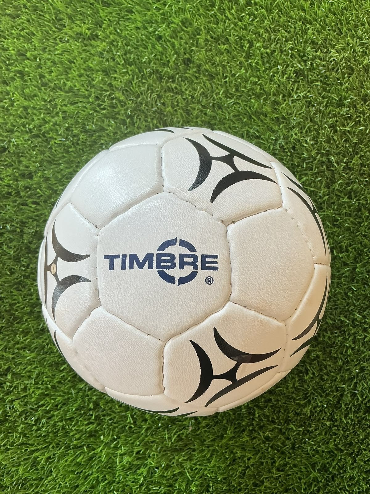 Bola Timbre branca e preta com logo azul sobre grama sintética